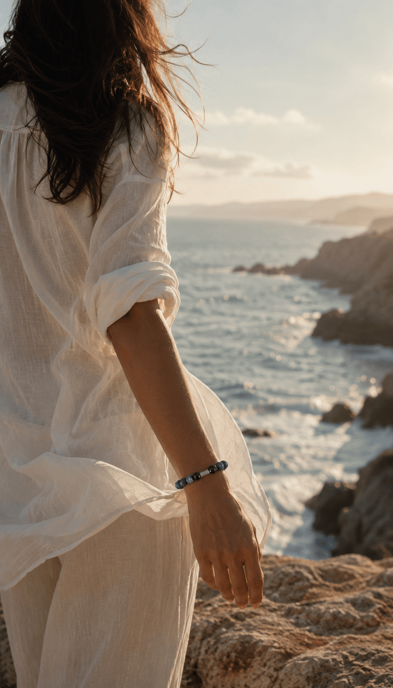 Woman in white linen standing at the edge of a rocky coastline, wearing a beaded bracelet